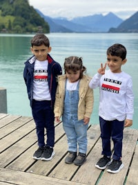 three children standing on a dock next to a lake